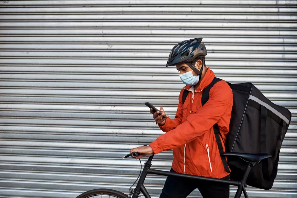 Man in orange jacket wearing surgical mask and bike helmet looks at phone while wearing delivery backpack and holding bicycle